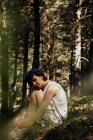 Vue latérale de romantique réfléchi jeune femme aux cheveux courts regardant la caméra en robe d'été et couronne florale embrassant les genoux tout en étant assis sur l'herbe dans la forêt luxuriante — Photo de stock
