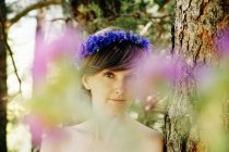 Peaceful adult female with bare shoulder and floral wreath on head standing near tree and looking at camera on sunny day in forest — Stock Photo