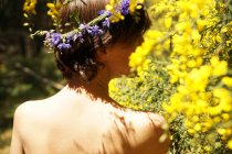 Обратный вид спокойной взрослой обнаженной женщины с цветочным венком на голове, отдыхающей в саду рядом с цветущим деревом с желтыми цветами в солнечный день — стоковое фото