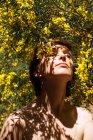 Низький кут спокійної дорослої гола жінка з закритими очима відпочиває в саду біля квітучого дерева з жовтими квітами в сонячний день — стокове фото