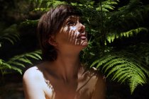 Alluring young naked woman with dark hair sitting near fern bush in lush tropical forest and looking up on sunny day — Stock Photo