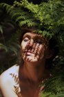 Alluring young naked woman with dark hair sitting near fern bush in lush tropical forest with eyes closed on sunny day — Stock Photo