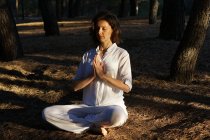 Side view of calm barefooted adult female in casual clothes meditating with closed eyes in Lotus asana while sitting on ground in forest on sunny day and practicing yoga — Stock Photo