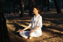 Вид збоку спокійного босоніж дорослої жінки в повсякденному одязі, роздумуючи із закритими очима в Lotus asana, сидячи на землі в лісі в сонячний день і практикуючи йогу — стокове фото