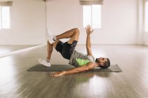 Side view of muscular young bearded male athlete lying on mat near mirror and doing Glute Bridge March exercise while training alone in spacious light studio — Stock Photo