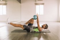 Side view of determined muscular young sportsman in activewear doing Leg Crunch exercise with medicine ball while lying on mat during workout in light studio — Stock Photo