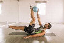 Вид збоку на визначеного м'язистого молодого спортсмена в активному одязі, що робить вправи ноги Крупп з медичним м'ячем, лежачи на килимку під час тренувань у легкій студії — стокове фото