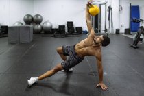 Poderoso muscular jovem atleta masculino com tronco nu em pé na prancha lateral e levantando kettlebell pesado durante o treino no ginásio — Fotografia de Stock