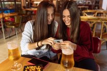 Молодая веселая лесбийская пара сидит за столом с бокалами пива в кафе и пользуется смартфоном, проводя выходные вместе — стоковое фото