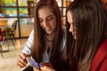 Молода весела лесбійська пара сидить за столом в кафе і використовує смартфон, проводячи вихідні разом — стокове фото