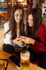 Jeune couple lesbien joyeux assis à table avec des verres de bière dans un café et en utilisant un smartphone tout en passant le week-end ensemble — Photo de stock
