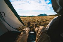 Crop unrecognizable male traveler in casual clothes lying in camping tent and admiring picturesque mountain landscape on sunny day — Stock Photo