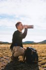 Side view of young stylish male traveler in casual outfit sitting on haunches near backpack and drinking hot beverage from thermos during hiking trip in mountainous terrain — Stock Photo