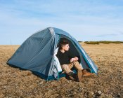 Full body of trendy young male traveler in stylish outfit sitting in camping tent while resting after trekking — Stock Photo