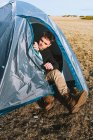 Corpo inteiro de jovem viajante masculino na moda em roupa elegante sentado na barraca de acampamento enquanto descansa após o trekking — Fotografia de Stock