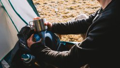 Side view of anonymous calm young male camper inc casual clothes enjoying hot drink while resting in tent after hiking in mountains on sunny day — Stock Photo