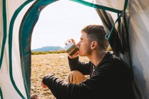 Caminhante masculino na moda jovem confiante em roupa quente beber caneca de bebida quente enquanto recria na barraca de acampamento no dia ensolarado — Fotografia de Stock