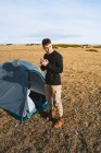 Selbstbewusster junger trendiger männlicher Wanderer im warmen Outfit trinkt einen Becher Heißgetränk und erholt sich am sonnigen Tag im Zeltlager — Stockfoto