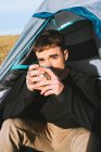 Caminhante masculino na moda jovem confiante em roupa quente caneca bebendo de bebida quente e olhando para a câmera enquanto recria na barraca de acampamento no dia ensolarado — Fotografia de Stock