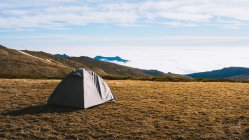Живописный пейзаж палатки для кемпинга, расположенной на травянистом склоне холма в заснеженной горной долине против облачного неба при солнечном свете — стоковое фото
