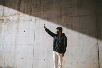 Unerkennbarer junger Mann in Kapuzenpulli und Maske steht auf der Straße in der Nähe einer Betonmauer und verdeckt das Gesicht mit der Hand vor grellem Sonnenlicht — Stockfoto
