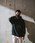 Модний хлопець у масці для обличчя, що регулює светр і дивиться геть, стоячи на вулиці біля стіни шерсті в сонячний день — стокове фото
