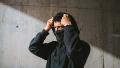 Anonymous trendy guy in face mask adjusting hoodie and looking away while standing on street near shabby wall on sunny day — Stock Photo