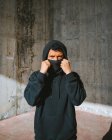 Unrecognizable young male in hoodie and mask standing on street near concrete wall — Stock Photo