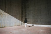 Full body of unrecognizable male in stylish hoodie and protective mask standing in abandoned building near shabby concrete walls and looking away — Stock Photo
