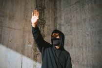 Unerkennbarer junger Mann in Kapuzenpulli und Maske steht auf der Straße in der Nähe einer Betonmauer und verdeckt das Gesicht mit der Hand vor grellem Sonnenlicht — Stockfoto