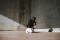 Irriconoscibile giovane maschio in felpa con cappuccio e maschera seduto sulla strada vicino al muro di cemento — Foto stock
