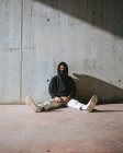 Unrecognizable young male in hoodie and mask sitting on street near concrete wall — Stock Photo