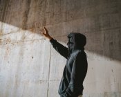 Unrecognizable young male in hoodie and mask standing on street near concrete wall and covering face with hand from bright sunlight — Stock Photo