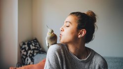 Молодая владелица в повседневной одежде готовится поцеловать очаровательную какатильскую птицу, сидящую на плече в гостиной — стоковое фото
