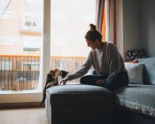 Side view of young lady in casual clothes sitting on comfortable couch with crossed legs and playing with adorable calico cat in modern apartment — Stock Photo