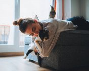 Вид збоку молодої леді в повсякденному одязі лежить на зручному дивані, граючи з чарівною каліко-кішкою в сучасній квартирі — стокове фото