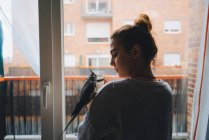 Симпатична пташка, що сидить на плечі молодої вдумливої жінки-власника в теплому светрі, стоїть біля вікна і п'є чашку гарячого напою вдома — стокове фото