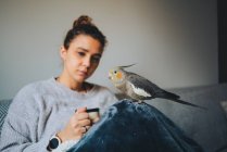 Junge Dame im warmen Pullover lächelt und trinkt heißen Kaffee, während sie es sich auf dem Sofa mit einem entzückenden Nymphenvogel auf der Hand gemütlich macht — Stockfoto