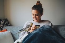 Щаслива молода леді в теплому светрі посміхається і п'є гарячу каву, відпочиваючи на дивані з чарівною пташкою на руці — стокове фото