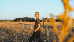Vista lateral de confiante jovem viajante feminino em roupas casuais e óculos em pé no prado e olhando para longe enquanto relaxa no campo ao pôr do sol — Fotografia de Stock
