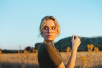 Vista lateral de confiante jovem viajante feminino em roupas casuais e óculos em pé no prado e olhando para longe enquanto relaxa no campo ao pôr do sol — Fotografia de Stock