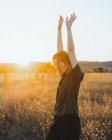 Seitenansicht einer jungen Frau in lässiger Kleidung, die den Sommertag genießt, während sie mit erhobenen Armen im trockenen Feld bei Sonnenuntergang in der Landschaft steht — Stockfoto