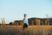 Vista lateral de la joven con el pelo rubio en ropa elegante caminando en medio de la hierba en el campo rural cerca de las colinas contra el cielo azul sin nubes al atardecer - foto de stock