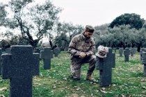 Full body sorrowful soldier in camouflage outfit kneeling down in front of grave in military cemetery on early autumn day — Stock Photo