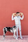 Full body of trendy young male in casual clothes holing skateboard behind head and looking away while standing on street with adorable Harlequin Great Dane dog — Stock Photo