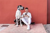 Полное тело счастливого молодого владельца мужчины, сидящего на скейтборде рядом с послушной собакой-датчанином Арлекин Грейт Дейн и общающегося по мобильному телефону на улице — стоковое фото