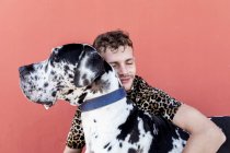 Vista laterale di felice giovane maschio unshaven in abiti casual e adorabile obbediente Arlecchino Grande Dane cane abbracciarsi su sfondo rosso — Foto stock