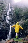 Back view of unrecognizable female teenager in bright raincoat with raised arms standing against cascade with fast water flow on mount — Stock Photo