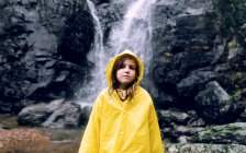 Female teenager in bright raincoat standing looking at camera against cascade with fast water flow on mount — Stock Photo