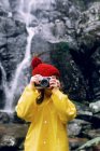 Adolescente anónimo en impermeable tomando fotos en cámara contra montaje en bruto con cascada espumosa a la luz del día - foto de stock